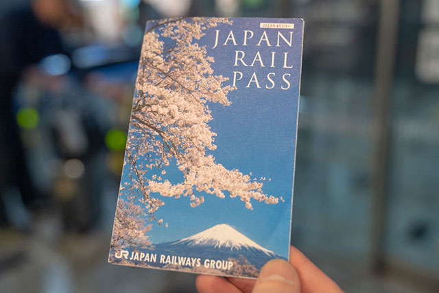 日本鉄道周遊券（JRパス）の実物チケット。背景には出発を待つ新幹線が写り、無制限の日本鉄道の旅を象徴している。